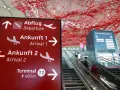 Czerwony znak z kierunkami na lotnisku w Niemczech: odloty, przyloty, terminal T2. W tle schody ruchome i instalacja artystyczna.