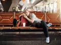 Zmęczona podróżniczka śpi na lotnisku, próbując pokonać jetlag. Obok niej plecak i kubek kawy.