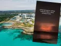 Widok na kurort na Cyprze. Na pierwszym planie widać turkusowe morze i plażę, a w tle białe budynki hotelowe. Na zdjęciu widnieje też napis: "Jesteś na Cyprze i widzisz wybuchy rakiet z plaży swojego hotelu...".