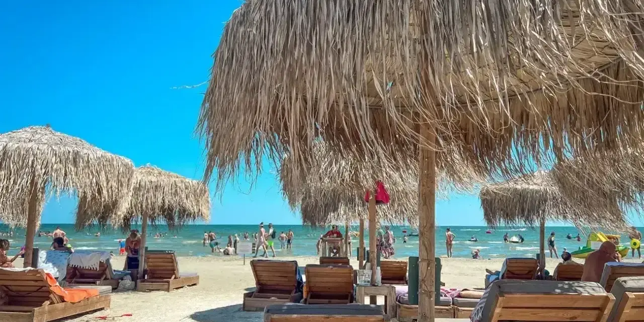 Słoneczne rumuńskie plaże z drewnianymi leżakami i strzechowymi parasolami. W tle widać ludzi kąpiących się w morzu.