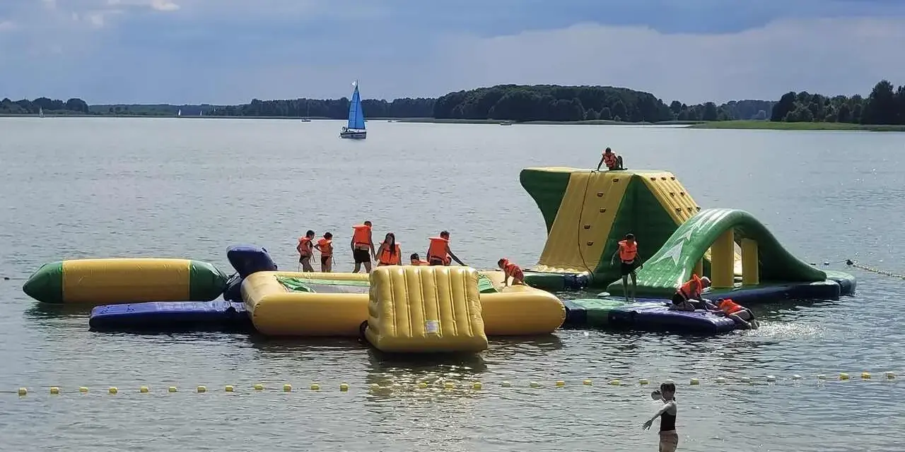 Dzieci w kamizelkach ratunkowych bawią się na dmuchanych wodnych atrakcjach. Idealne miejsce, gdzie na wakacje z dziećmi, z żaglówką na horyzoncie.