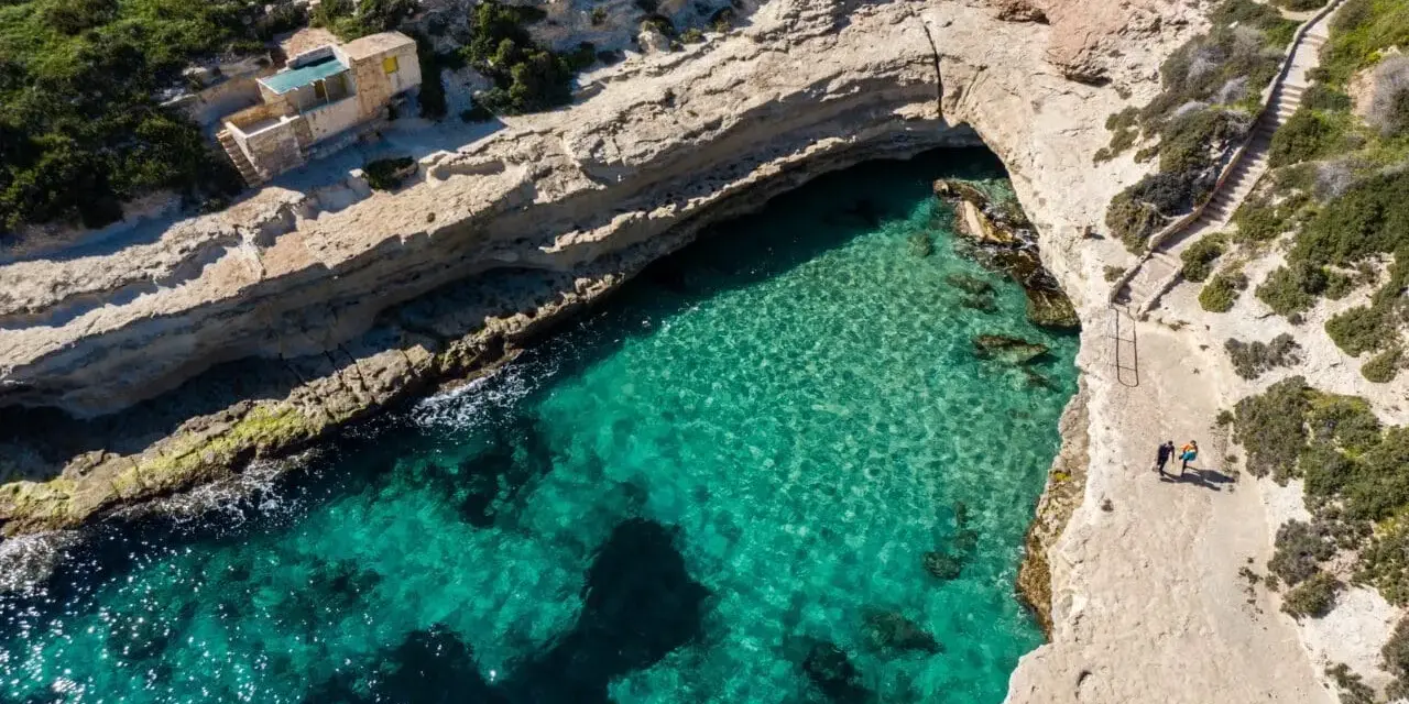 Malta plaże: turkusowa woda w zatoce, skaliste wybrzeże i schody prowadzące do ukrytej plaży. Dwie osoby spacerują po klifie.
