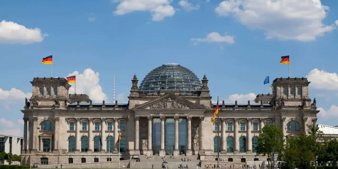 Imponujący Reichstag z charakterystyczną szklaną kopułą, otoczony zielonym trawnikiem i błękitnym niebem z chmurami.