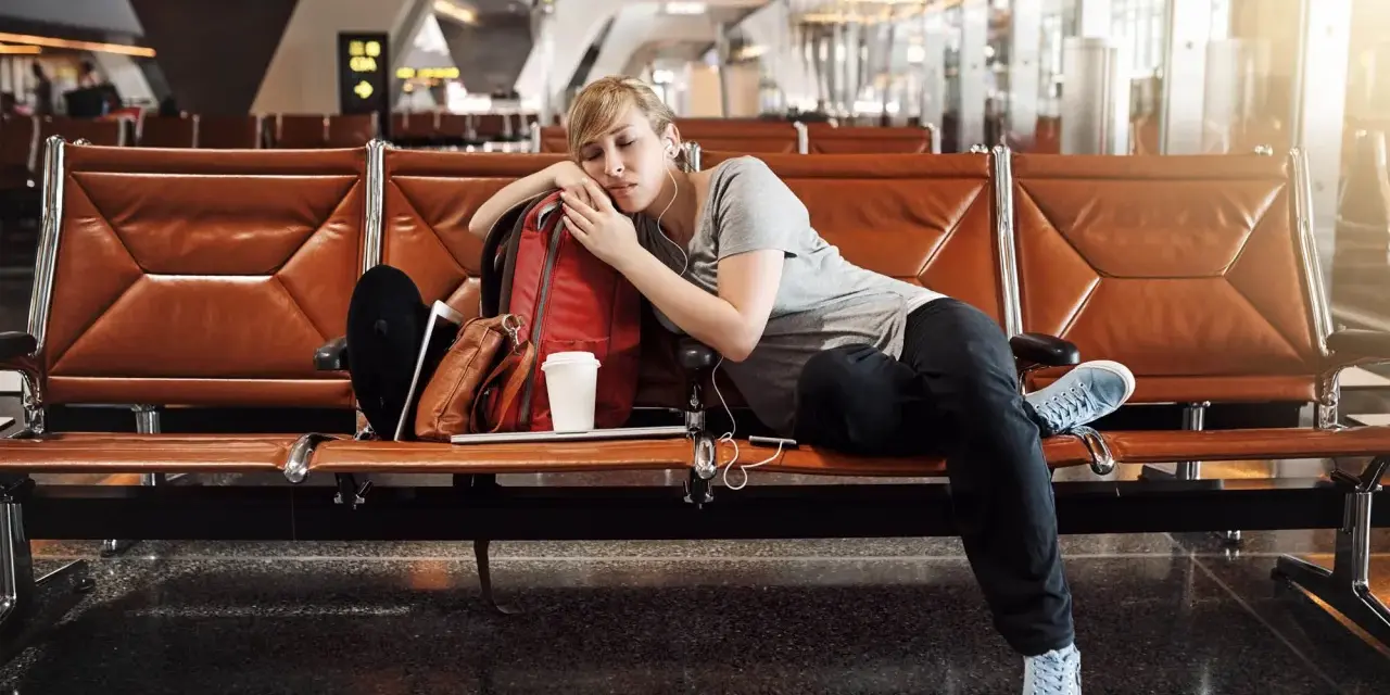 Zmęczona podróżniczka śpi na lotnisku, próbując pokonać jetlag. Obok niej plecak i kubek kawy.
