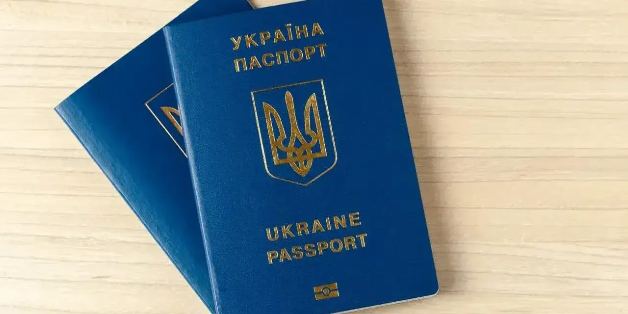 Dwa niebieskie paszporty biometryczne Ukrainy z złotym godłem i napisami leżą na drewnianym blacie.
