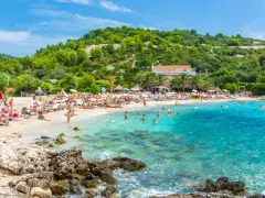 Słoneczna plaża z turkusową wodą, mnóstwem ludzi odpoczywających na leżakach i kąpiących się w morzu. W tle zielone wzgórza i budynek z czerwoną dachem. Idealne miejsce na pierwszy minutę wakacji.