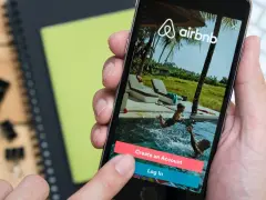 Dłoń trzyma smartfon z aplikacją Airbnb. Czy Airbnb jest bezpieczne? Na ekranie widać basen i dzieci, przyciski "Create an Account" i "Log In".