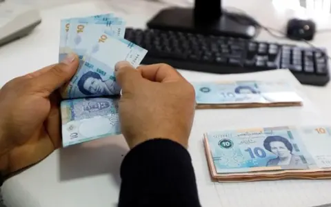 Ręce liczą banknoty 10 dinarów, tunezyjska waluta. Na stole leżą stosy pieniędzy, a w tle widać klawiaturę komputera.