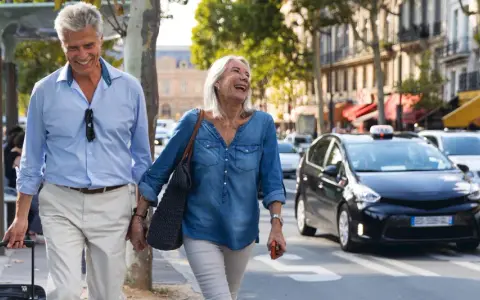 Uśmiechnięta para seniorów spaceruje po mieście z walizką, obok przejeżdżają samochody.