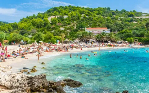 Słoneczna plaża z turkusową wodą, mnóstwem ludzi odpoczywających na leżakach i kąpiących się w morzu. W tle zielone wzgórza i budynek z czerwoną dachem. Idealne miejsce na pierwszy minutę wakacji.