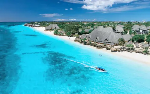 Widok na turkusowe morze i białą plażę z hotelami, kiedy na Zanzibarze. Łódź motorowa z pasażerami płynie po wodzie.