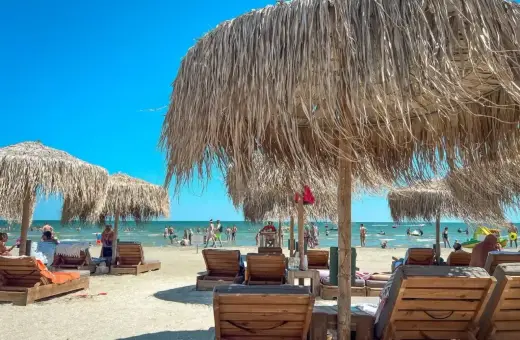 Słoneczne rumuńskie plaże z drewnianymi leżakami i strzechowymi parasolami. W tle widać ludzi kąpiących się w morzu.