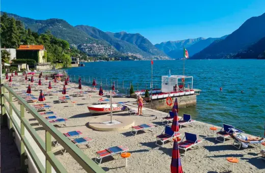 Plaże nad jeziorem Como, z rzędami leżaków i parasoli, zapraszają do relaksu. W tle góry i błękitne niebo.
