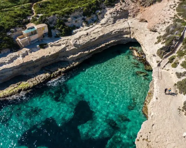 Malta plaże: turkusowa woda w zatoce, skaliste wybrzeże i schody prowadzące do ukrytej plaży. Dwie osoby spacerują po klifie.