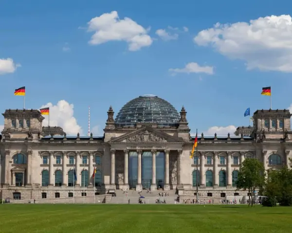 Imponujący Reichstag z charakterystyczną szklaną kopułą, otoczony zielonym trawnikiem i błękitnym niebem z chmurami.
