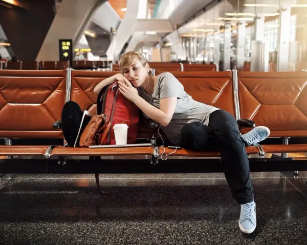 Zmęczona podróżniczka śpi na lotnisku, próbując pokonać jetlag. Obok niej plecak i kubek kawy.