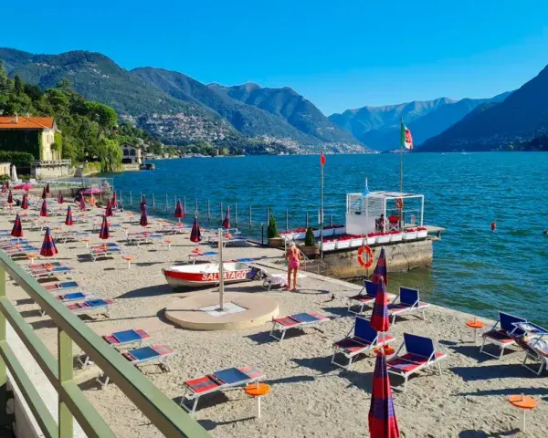 Plaże nad jeziorem Como, z rzędami leżaków i parasoli, zapraszają do relaksu. W tle góry i błękitne niebo.