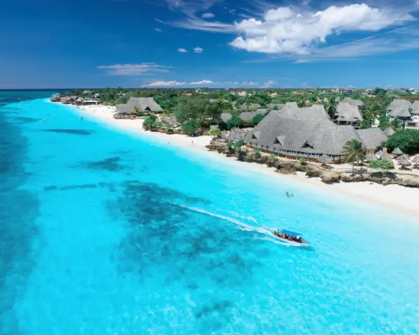 Widok na turkusowe morze i białą plażę z hotelami, kiedy na Zanzibarze. Łódź motorowa z pasażerami płynie po wodzie.