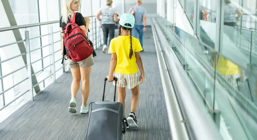 Młoda kobieta z plecakiem i dziewczynka z walizką idą korytarzem lotniska. To może być ich wyjazd nieletniego za granicę bez rodziców.