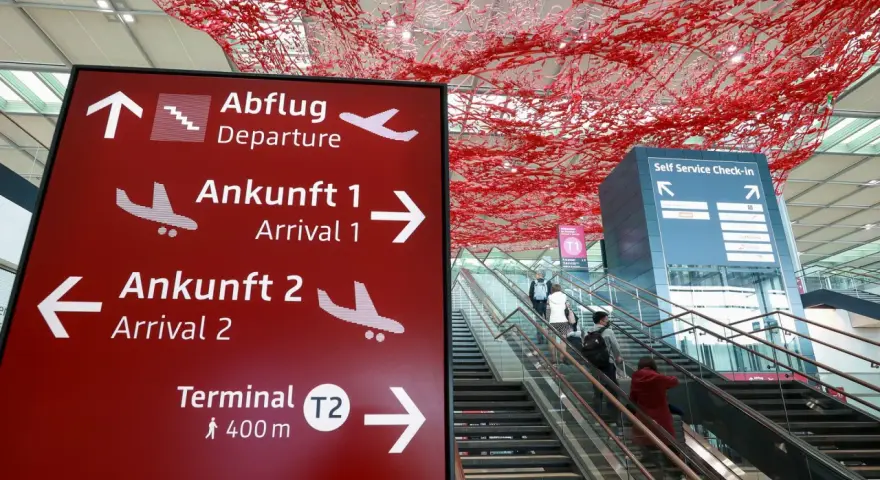 Czerwony znak z kierunkami na lotnisku w Niemczech: odloty, przyloty, terminal T2. W tle schody ruchome i instalacja artystyczna.