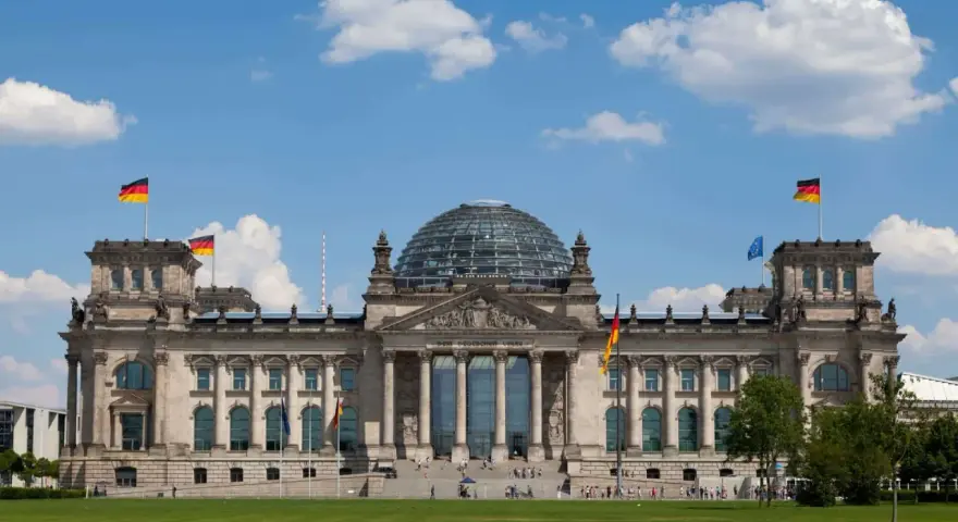 Imponujący Reichstag z charakterystyczną szklaną kopułą, otoczony zielonym trawnikiem i błękitnym niebem z chmurami.