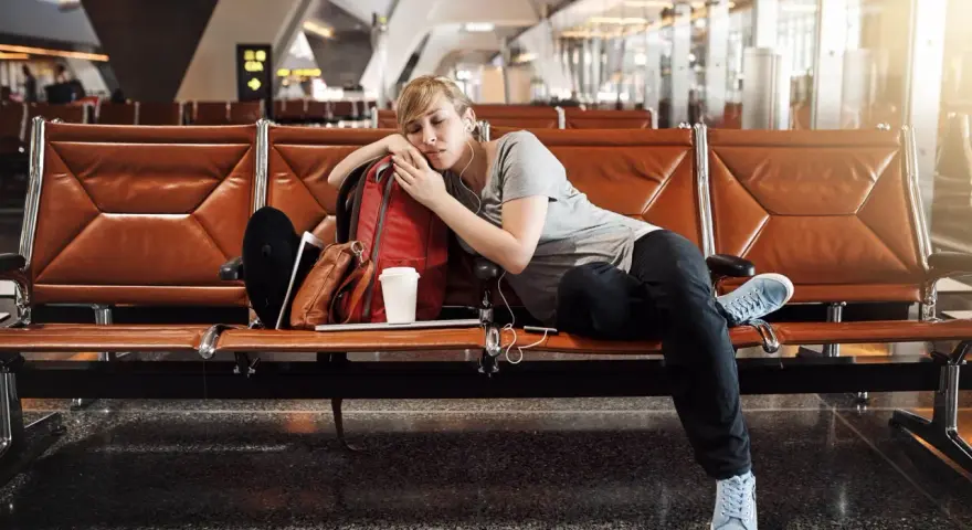 Zmęczona podróżniczka śpi na lotnisku, próbując pokonać jetlag. Obok niej plecak i kubek kawy.