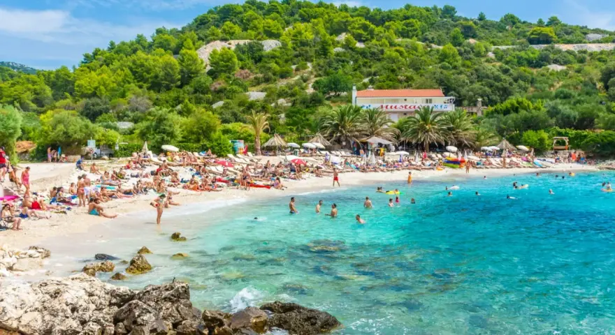 Słoneczna plaża z turkusową wodą, mnóstwem ludzi odpoczywających na leżakach i kąpiących się w morzu. W tle zielone wzgórza i budynek z czerwoną dachem. Idealne miejsce na pierwszy minutę wakacji.