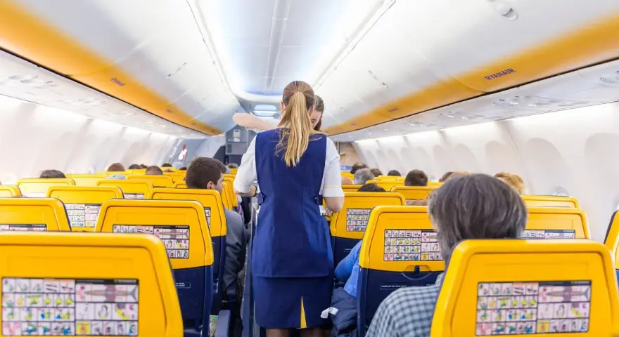 Wnętrze samolotu Ryanair, stewardessa obsługuje pasażerów. Żółte zagłówki siedzeń i pomarańczowe pasy na suficie tworzą słoneczną atmosferę.