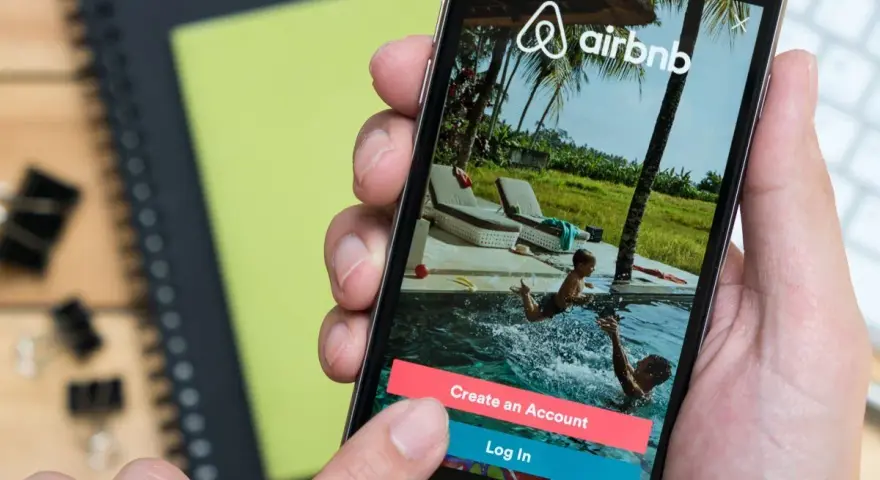 Dłoń trzyma smartfon z aplikacją Airbnb. Czy Airbnb jest bezpieczne? Na ekranie widać basen i dzieci, przyciski "Create an Account" i "Log In".