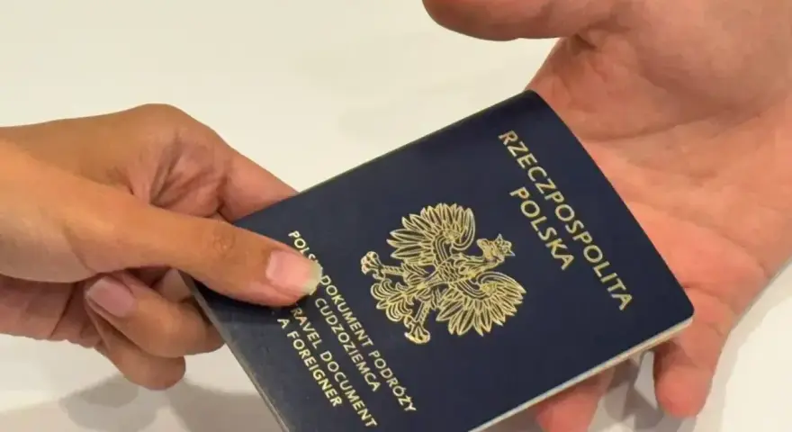 Dłoń przekazuje ciemnoniebieski paszport polski z godłem orła. Dokument podróży gotowy do przekroczenia granicy.