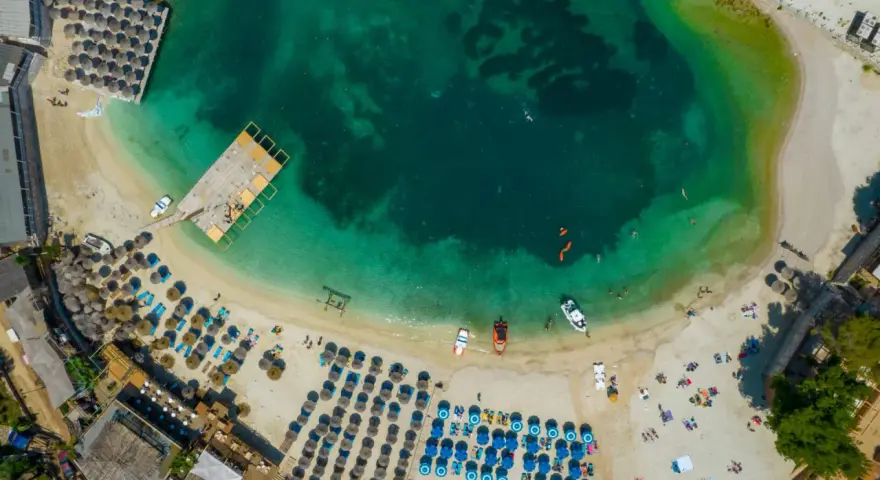 Widok z lotu ptaka na turkusowe morze i piaszczyste **albania plaże** z leżakami, parasolami i ludźmi cieszącymi się słońcem.