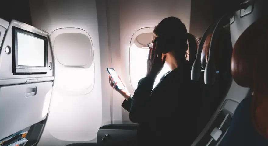 Kobieta w okularach trzyma telefon, zastanawiając się, czy w samolocie jest wifi. Obok ekran rozrywki.