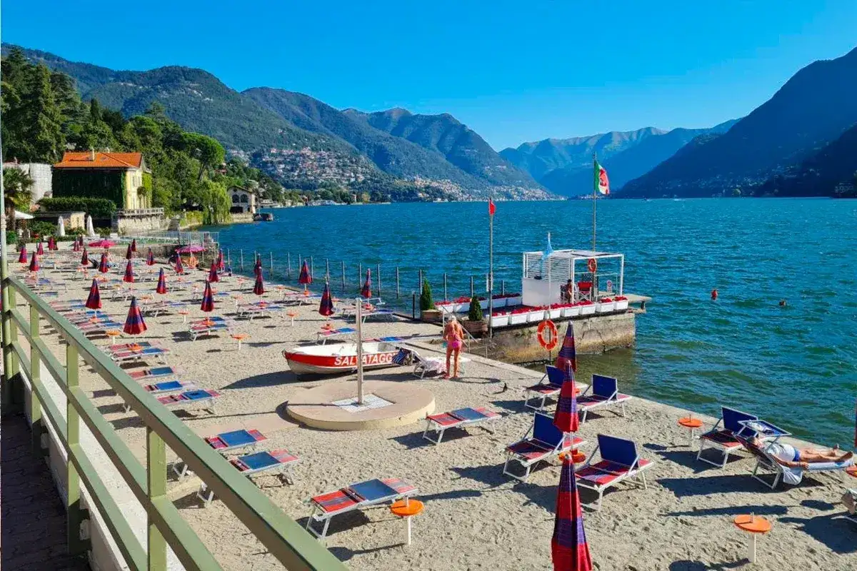 Plaże nad jeziorem Como, z rzędami leżaków i parasoli, zapraszają do relaksu. W tle góry i błękitne niebo.