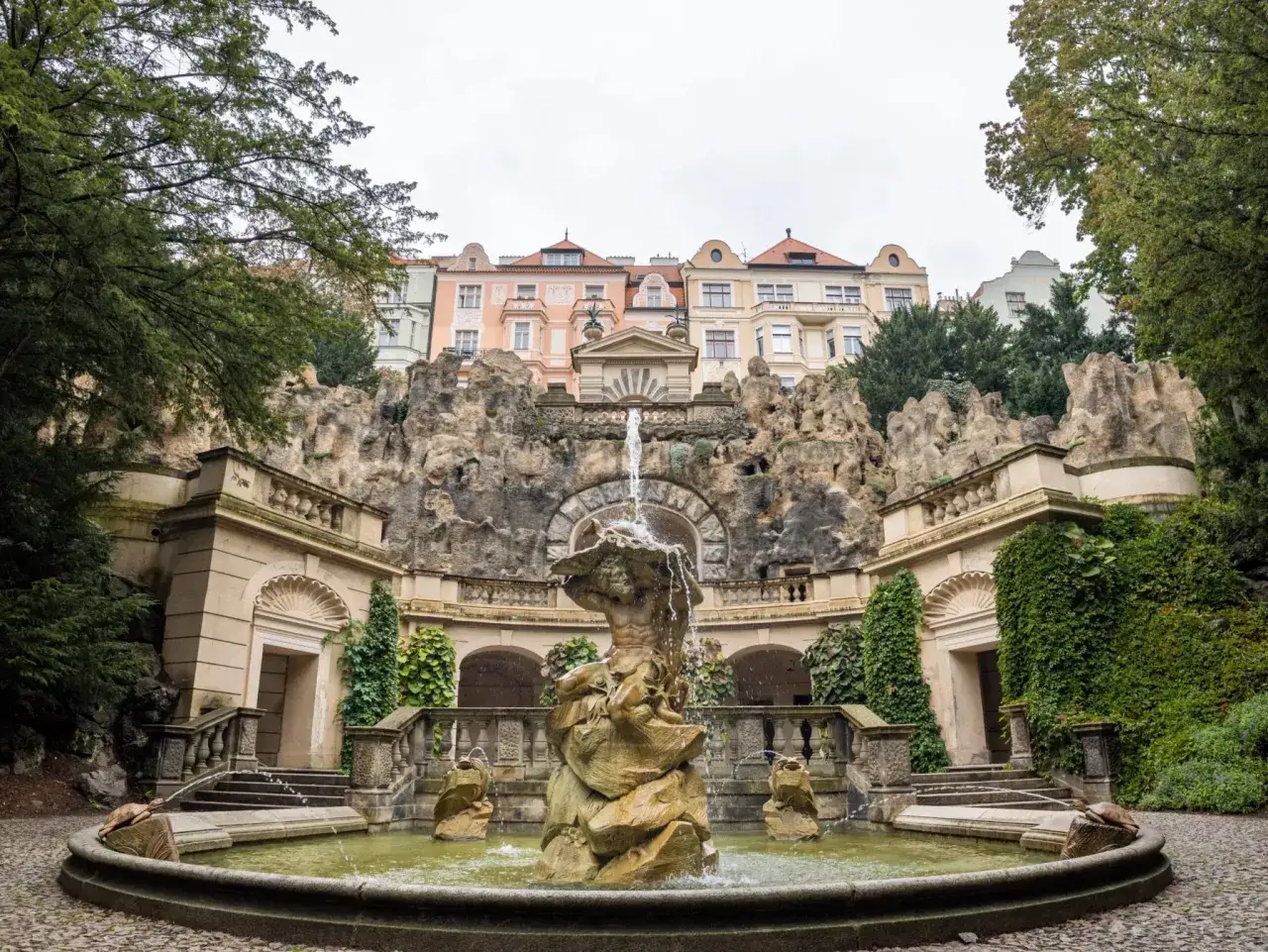Praga nietypowe atrakcje: fontanna z Atlantem w ogrodzie, ukryta wśród skał i zieleni, z barwnymi kamienicami w tle.