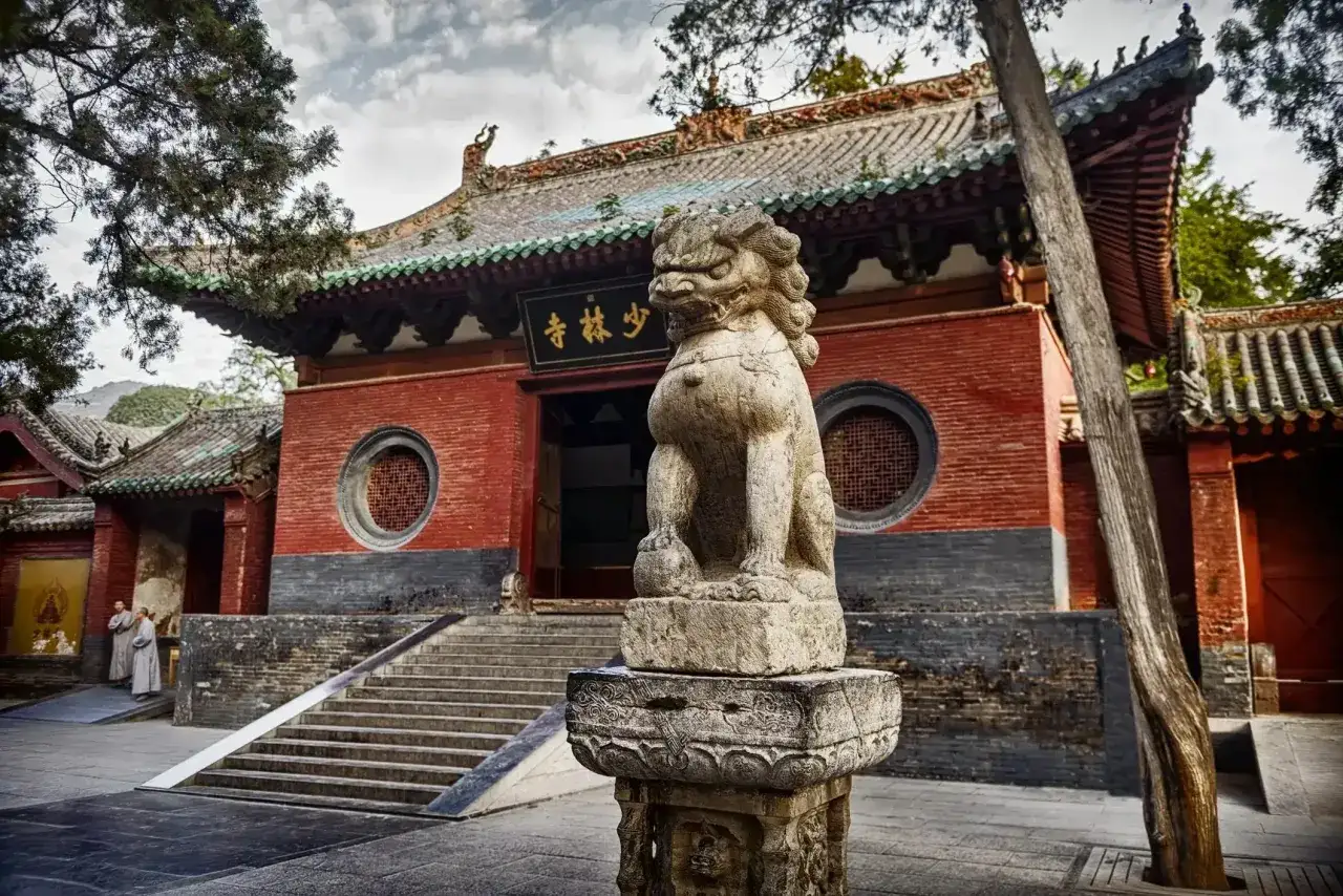 Kamienna lwica strzeże wejścia do klasztoru Shaolin. Dwie postacie w szatach mnichów stoją na schodach.