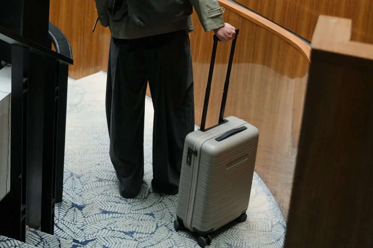 Mężczyzna ciągnie szarą walizkę, idealną jako bagaż podręczny Ryanair, po dywanie z wzorem liści.