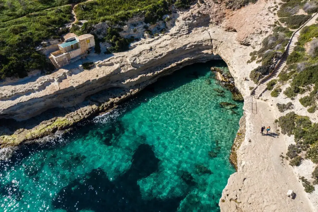Malta plaże: turkusowa woda w zatoce, skaliste wybrzeże, mały domek i ścieżka z dwoma spacerującymi osobami.