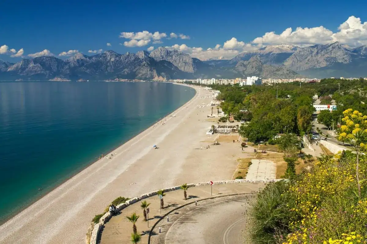 Słoneczny dzień na plaży Lara, z turkusowym morzem, piaszczystym brzegiem i górami w tle.