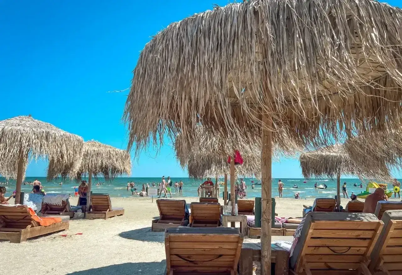 Słoneczne rumuńskie plaże z drewnianymi leżakami i strzechowymi parasolami. W tle widać ludzi kąpiących się w morzu.